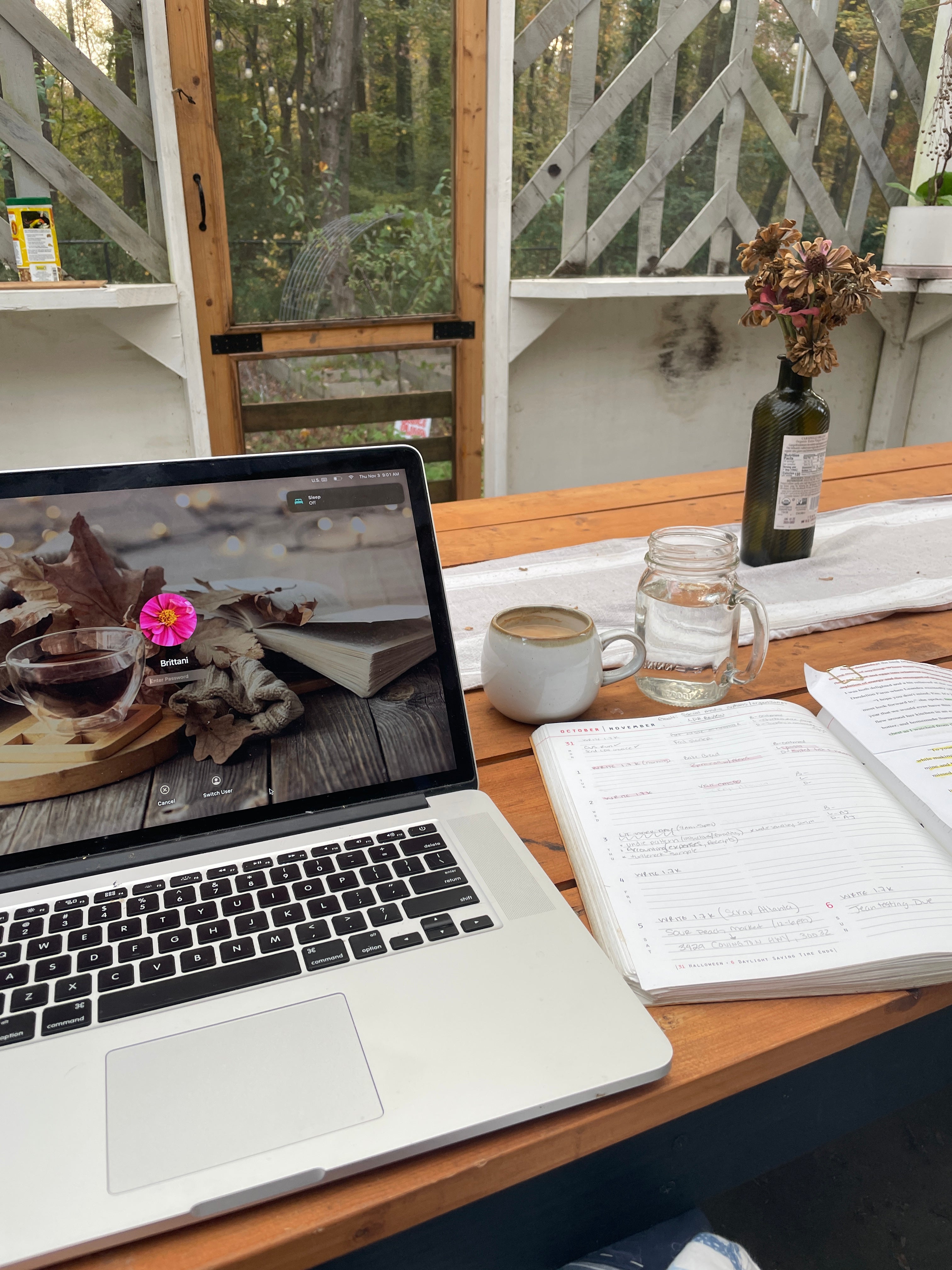 On a wooden table sits an open laptop with a fall inspired background including a glass cup of tea, cozy knitted blankets, and an open book. Next to the laptop sits a cream colored cup of coffee, a mason jar glass of water, and a planner spread open.