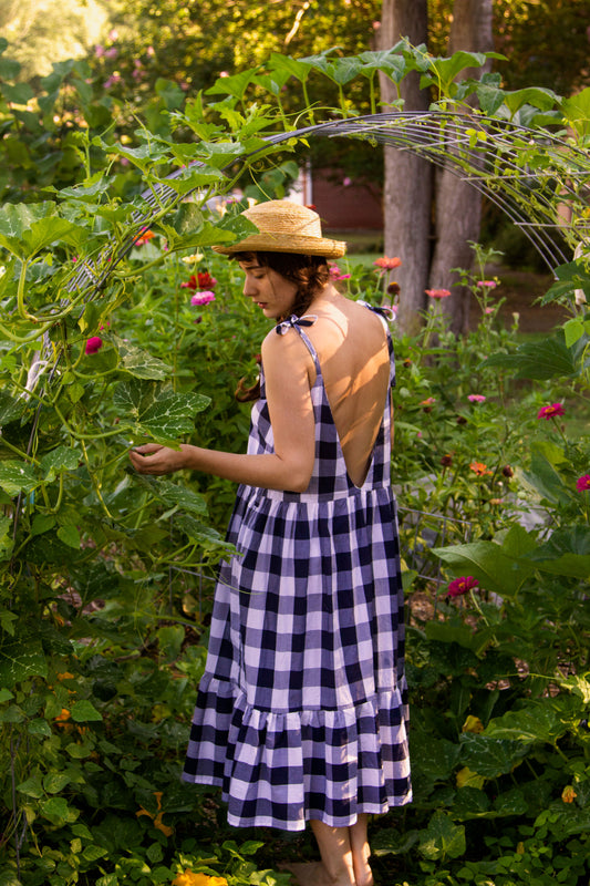 A white woman with long brown hair put into pigtail braids wears a straw hat and a blue gingham dress while standing under a garden arch