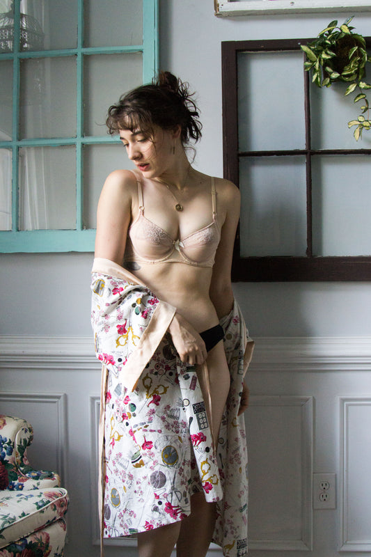 Brunette girl standing in front of window paned wall in a nude bra and a white floral robe hanging off her arms. 
