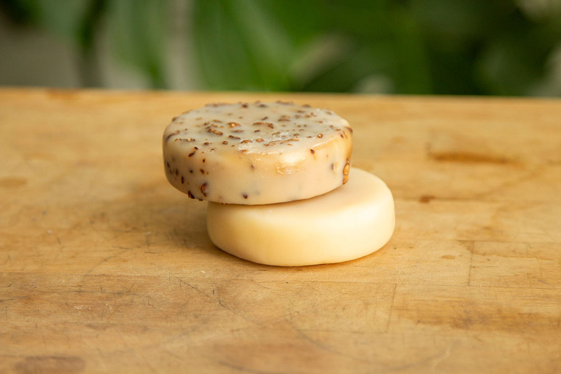 A stack of two circular bars of soap sitting on a light wooden desk with greenery in the background. The bottom bar of soap is a light beige color and the top bar is a darker beige with flecks of brown oatmeal throughout