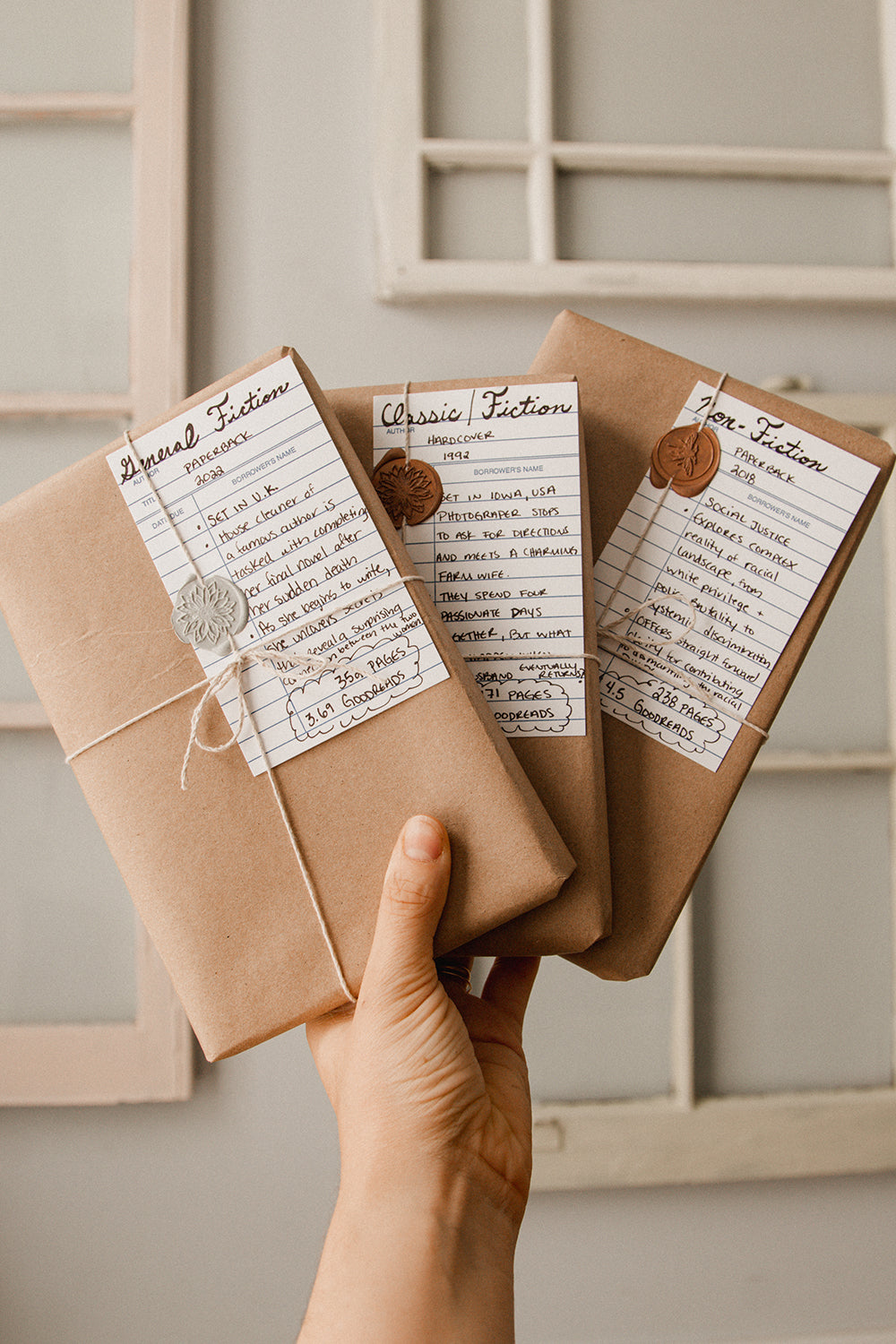 A white hand holds up three paper wrapped books, each with a twine bow tie, wax seal, and library card with handwritten descriptions.