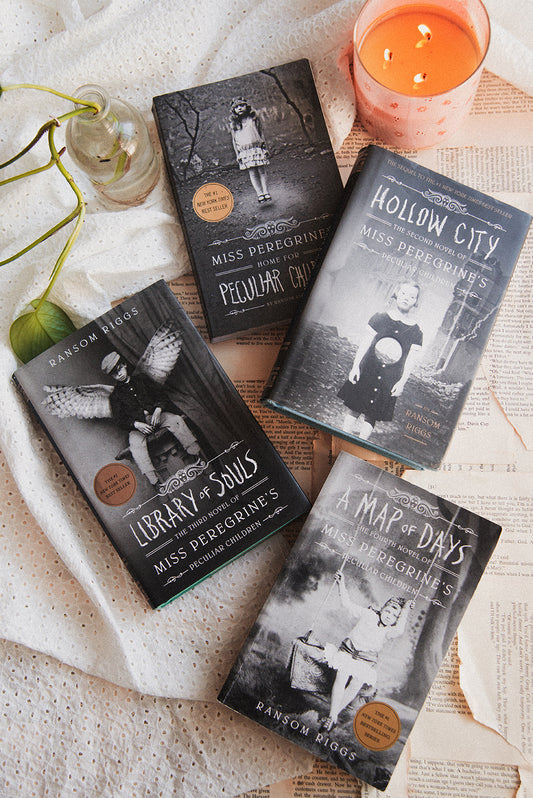 Four books by Miss Peregrine's Peculiar Children series on a textured surface with a candle and greenery.